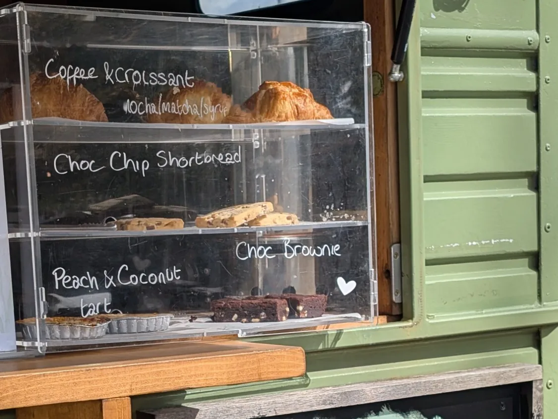 The bakes cabinet in the horsebox hatch at Bulford, holding croissants, chocolate-chip shortbread, brownies and a peach and coconut tart, each labelled by hand on the glass