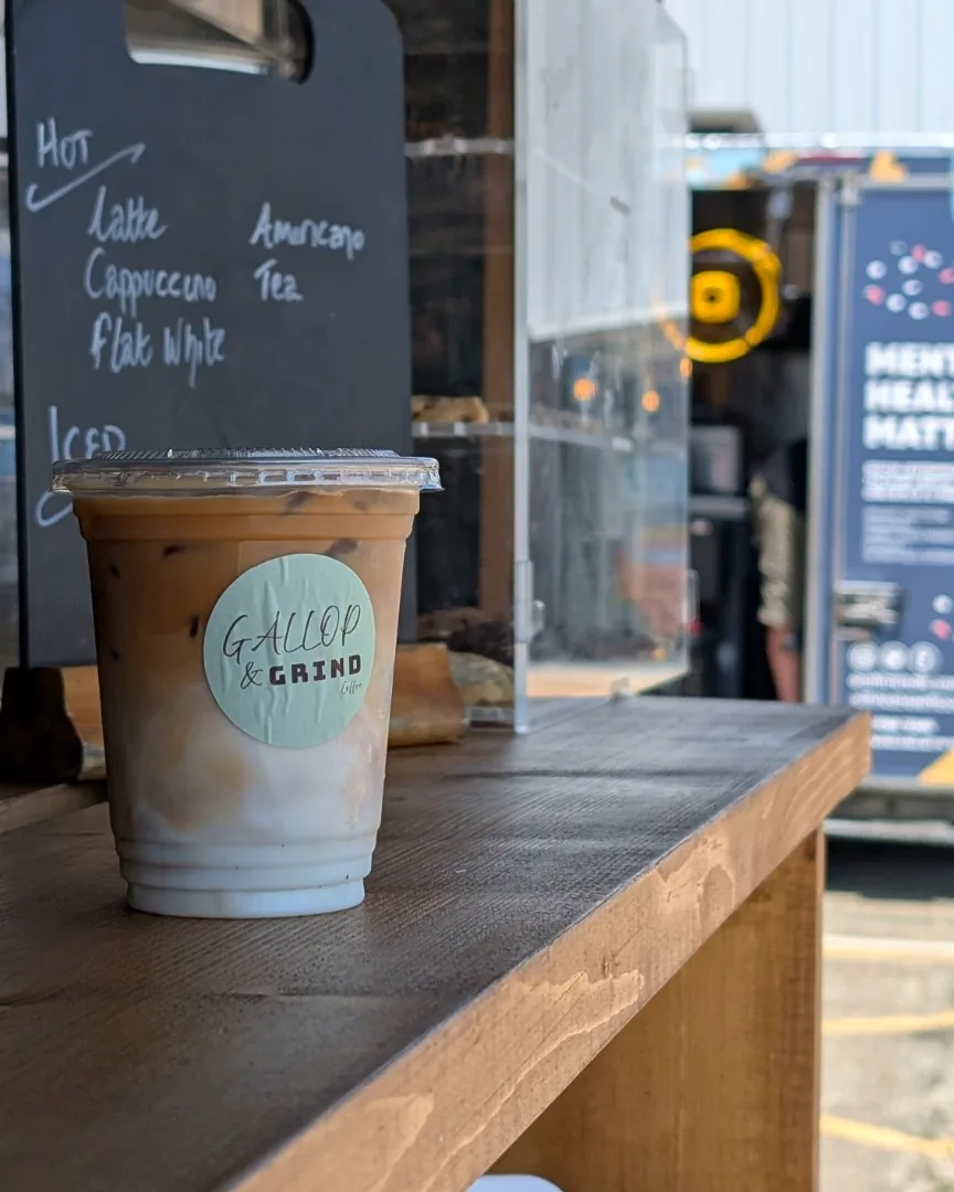 An iced latte in a branded Gallop & Grind cup on the wooden serving counter, with the hot and iced chalkboard menu behind