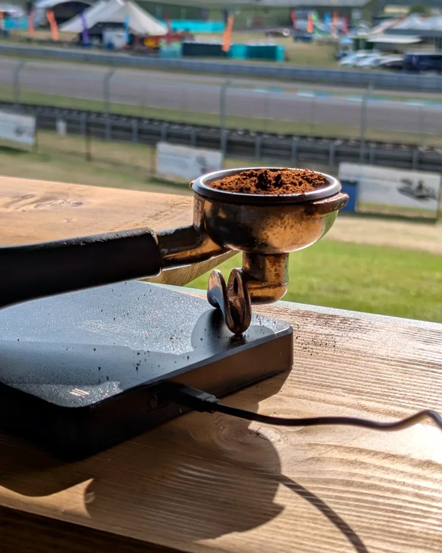 A dosed portafilter resting on the scales at the horsebox serving hatch, with an outdoor event ground and trade stands behind