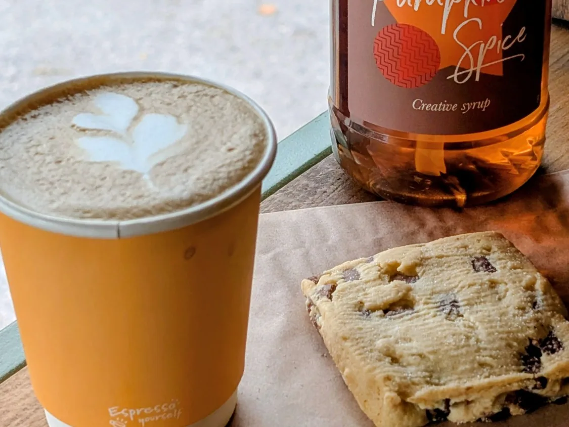 A seasonal spiced latte with heart latte art beside a bottle of pumpkin spice syrup and a chocolate-chip shortbread on the horsebox hatch ledge