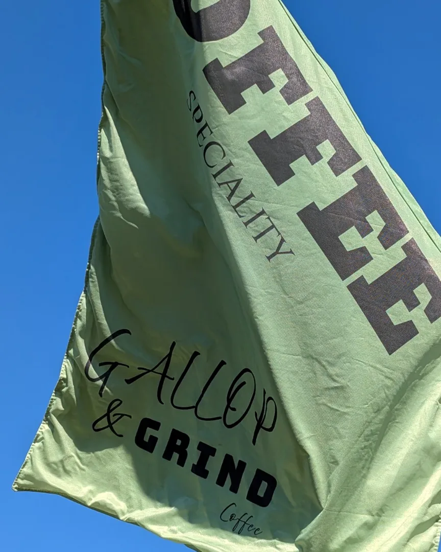 A sage-green Gallop & Grind speciality coffee flag flying against a clear blue sky above the event pitch